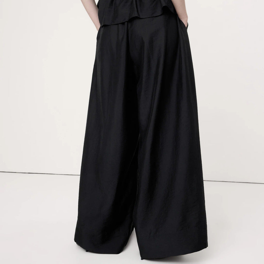 A model shown from the back wearing a black strapless jumpsuit with a ruched smocked bodice and very wide-leg pants, set against a plain off-white studio background.