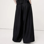 A model shown from the back wearing a black strapless jumpsuit with a ruched smocked bodice and very wide-leg pants, set against a plain off-white studio background.