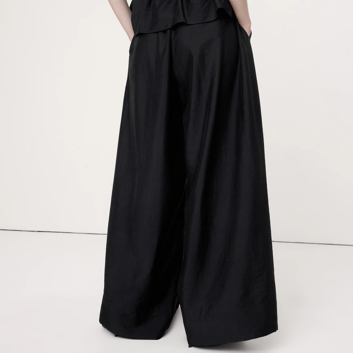 A model shown from the back wearing a black strapless jumpsuit with a ruched smocked bodice and very wide-leg pants, set against a plain off-white studio background.