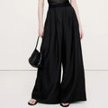 A model wearing a black sheer short-sleeve top with a structured bust and high-waisted, wide-leg black trousers, holding a black woven handbag against a plain white studio background.