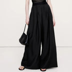 A model wearing a black sheer short-sleeve top with a structured bust and high-waisted, wide-leg black trousers, holding a black woven handbag against a plain white studio background.