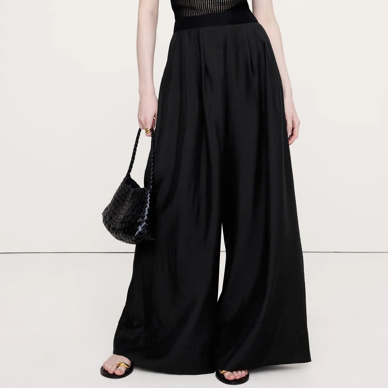 A model wearing a black sheer short-sleeve top with a structured bust and high-waisted, wide-leg black trousers, holding a black woven handbag against a plain white studio background.