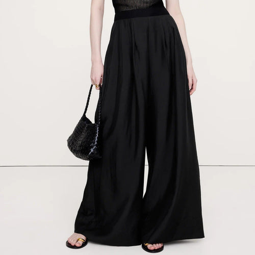 A model wearing a black sheer short-sleeve top with a structured bust and high-waisted, wide-leg black trousers, holding a black woven handbag against a plain white studio background.