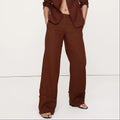 A model with curly dark hair wearing a matching rust-brown linen-blend two-piece suit, featuring a cropped blazer and wide-leg trousers, standing against a plain white studio background.