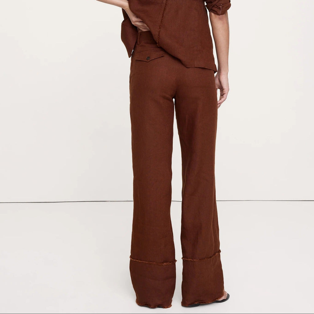 A model from behind wearing a matching brown linen-blend two-piece set consisting of a cropped long-sleeve top and wide-leg trousers, set against a plain white studio background.