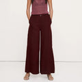 A woman modeling a sleeveless maroon knit top with a tie neckline and dark burgundy wide-leg trousers, standing against a plain white studio background.
