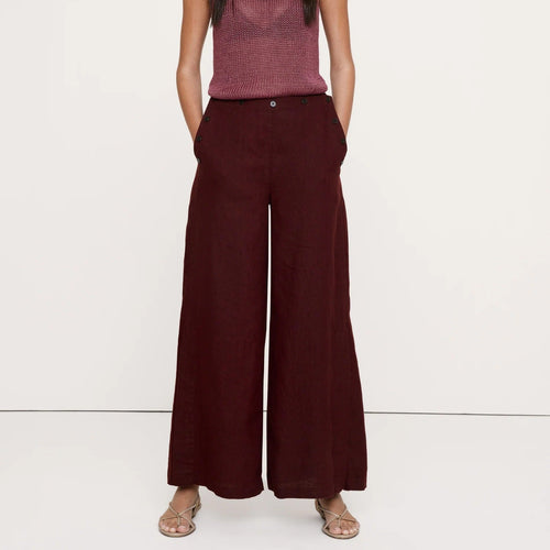 A woman modeling a sleeveless maroon knit top with a tie neckline and dark burgundy wide-leg trousers, standing against a plain white studio background.