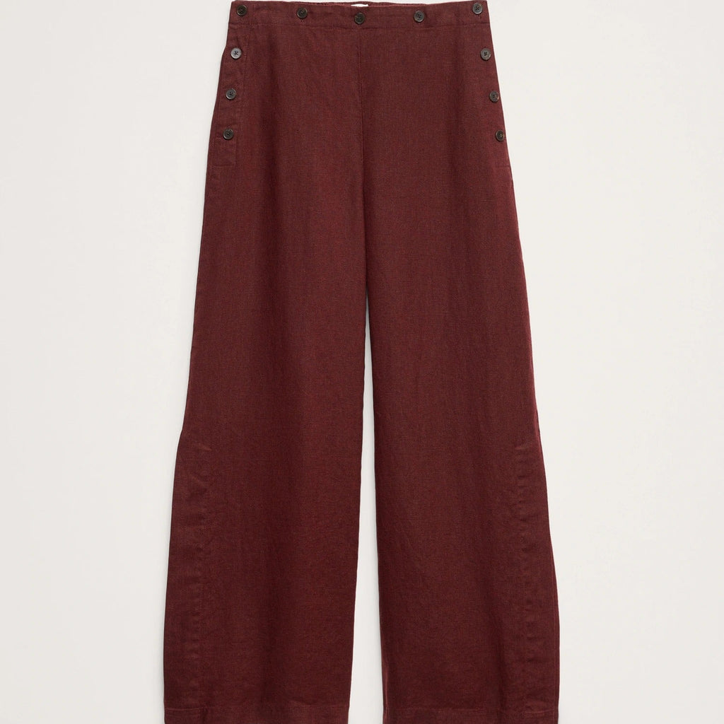 A pair of wide-leg trousers in a deep burgundy red, featuring decorative dark buttons along the outer seams of the legs, displayed flat against a light off-white background.