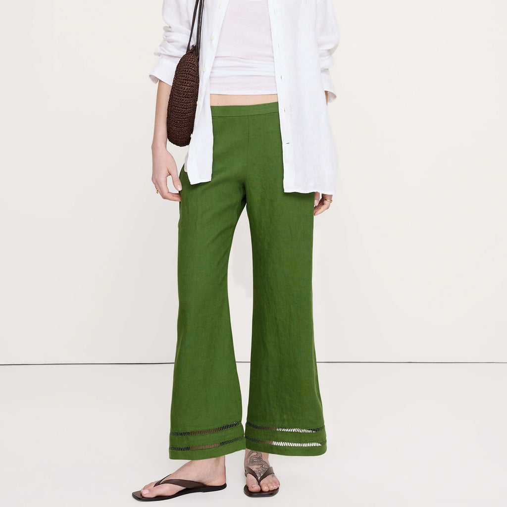 A woman modeling a pair of olive green wide-leg linen pants, styled with a white button-down shirt, white tank top, brown woven shoulder bag, and black flip-flops against a plain off-white studio background.