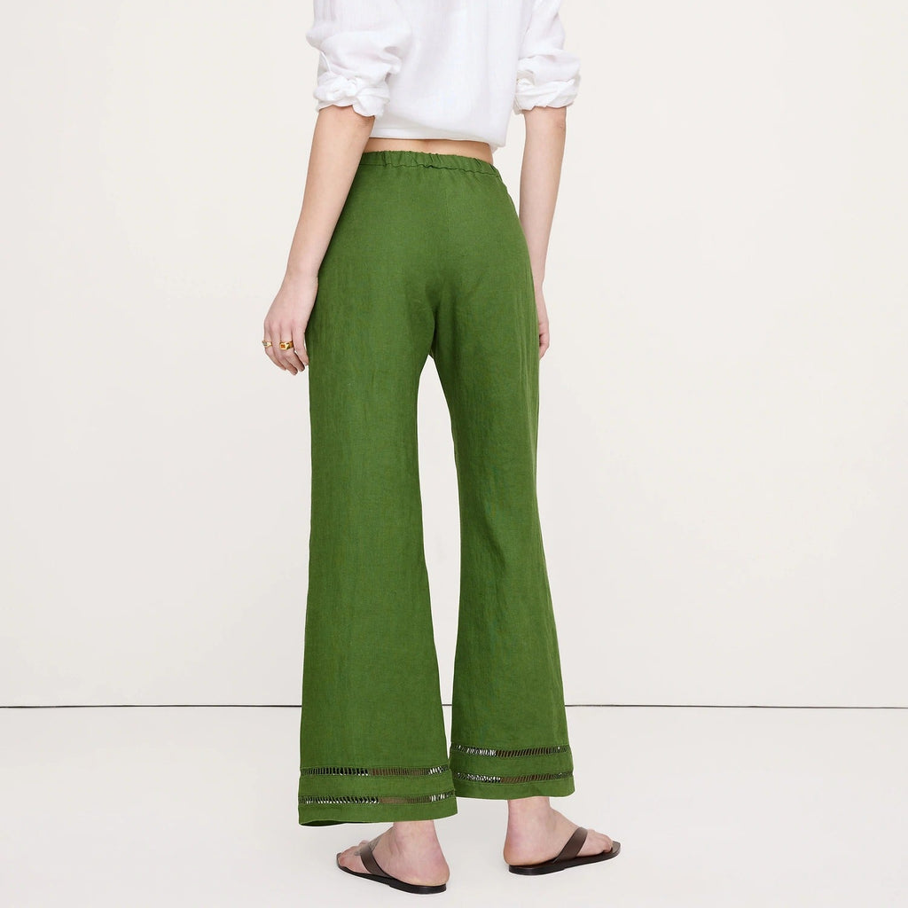 A woman shown from the back wearing olive green wide-leg trousers with a white blouse and black sandals, standing against a plain off-white studio background.