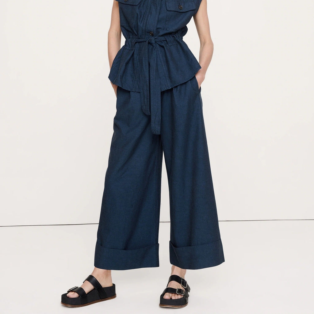 A woman modeling a dark navy blue sleeveless belted jumpsuit with wide legs and a high mock neck, paired with black sandals, against a plain white studio background.