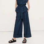 A woman modeling a dark navy blue sleeveless belted jumpsuit with wide legs and a high mock neck, paired with black sandals, against a plain white studio background.
