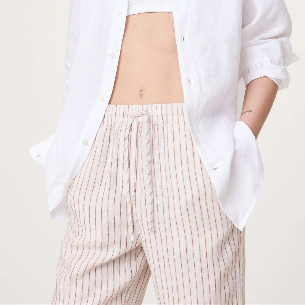 A model wearing a white linen button-up shirt over a white crop top, paired with beige and white vertically striped trousers, against a plain off-white background.