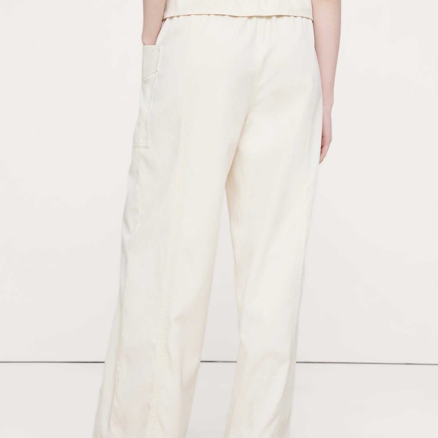 A person shown from the back wearing off-white wide-leg trousers with a gathered elastic waistband and side pockets, paired with a white ribbed top and black sandals, against a plain light background.