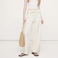A model stands against a plain off-white studio background wearing a white ribbed tank top, cream wide-leg drawstring pants, and black flip-flops, while holding a woven straw tote bag.