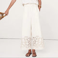 A woman modeling a white two-piece linen set featuring a short-sleeve top with floral embroidery and matching wide-leg trousers with lace detailing at the hem, set against a plain white studio background.