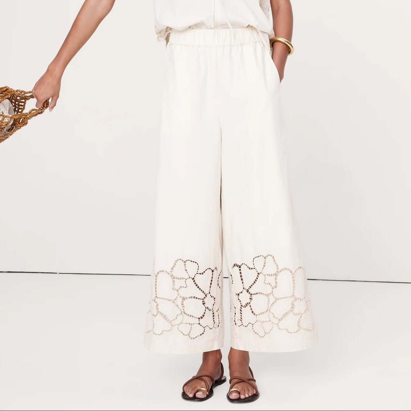 A woman modeling a white two-piece linen set featuring a short-sleeve top with floral embroidery and matching wide-leg trousers with lace detailing at the hem, set against a plain white studio background.