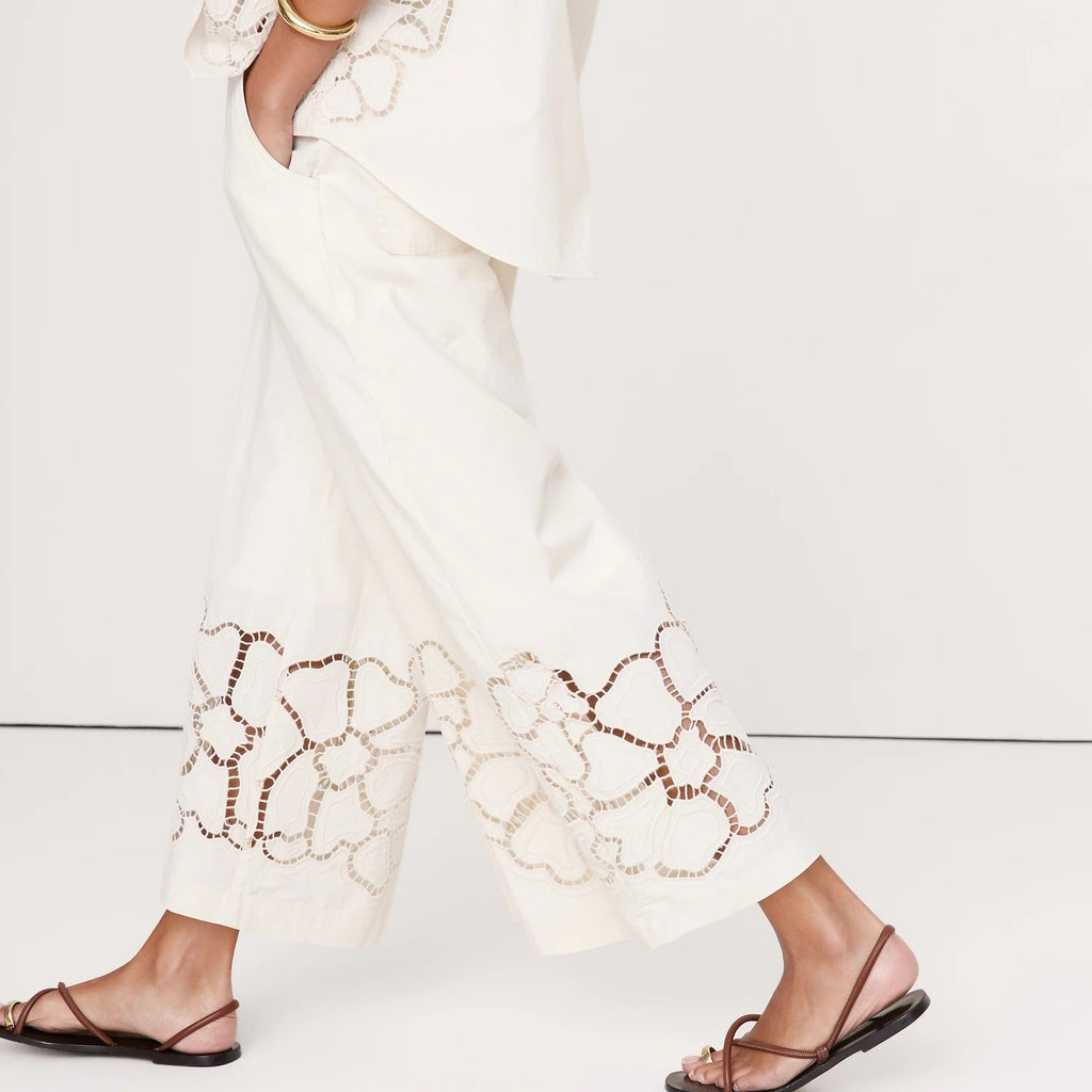 A model wearing a white sleeveless top and matching wide-leg cropped pants with intricate floral lace-up embroidery, styled with brown strappy flat sandals against a clean white studio background.